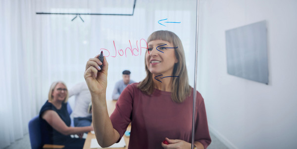 Eine Person steht hinter einer Glasscheibe und schreibt mit einem Filzstift Wörter auf das Glas. Im Hintergrund sitzen Personen an einem Konferenz-Tisch.